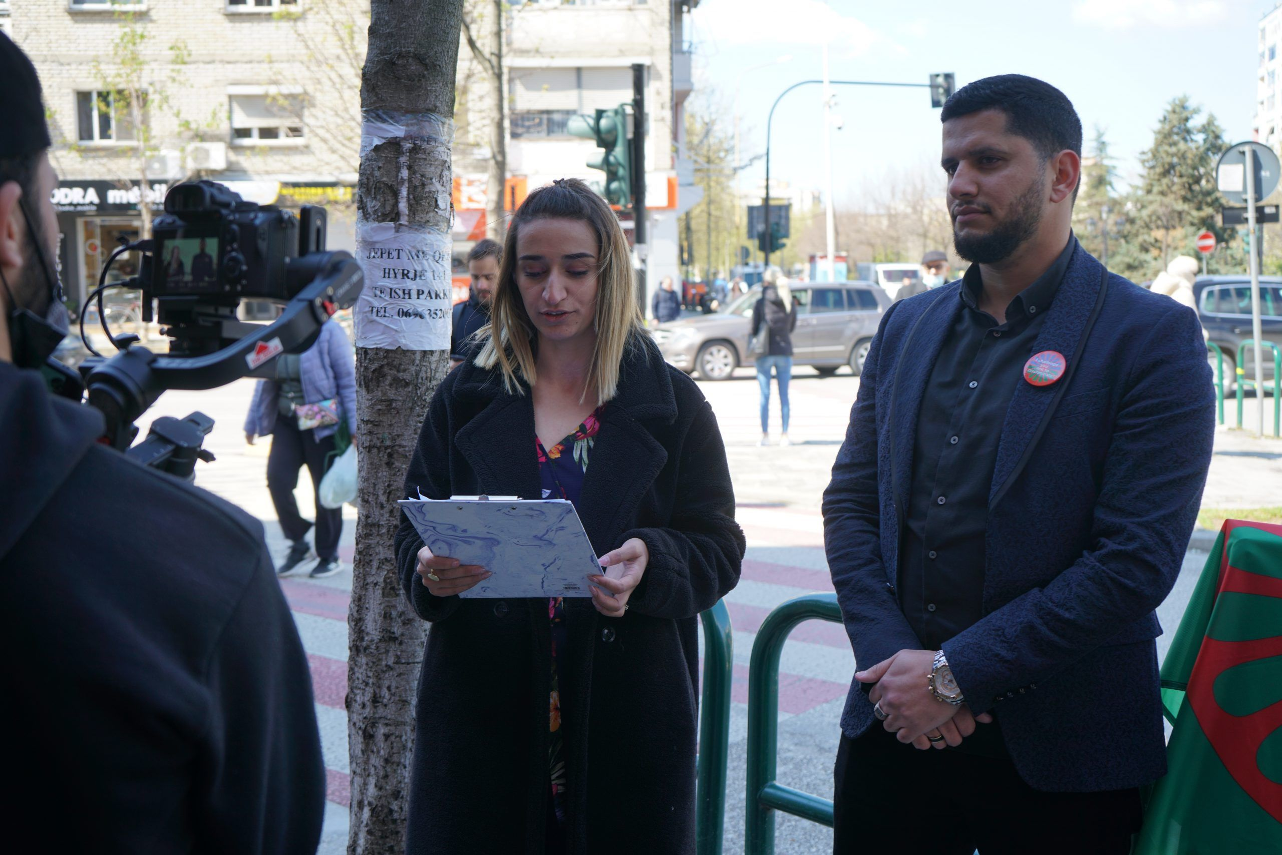 A speaker reading a statement at an outdoor commemoration while a camera crew films
