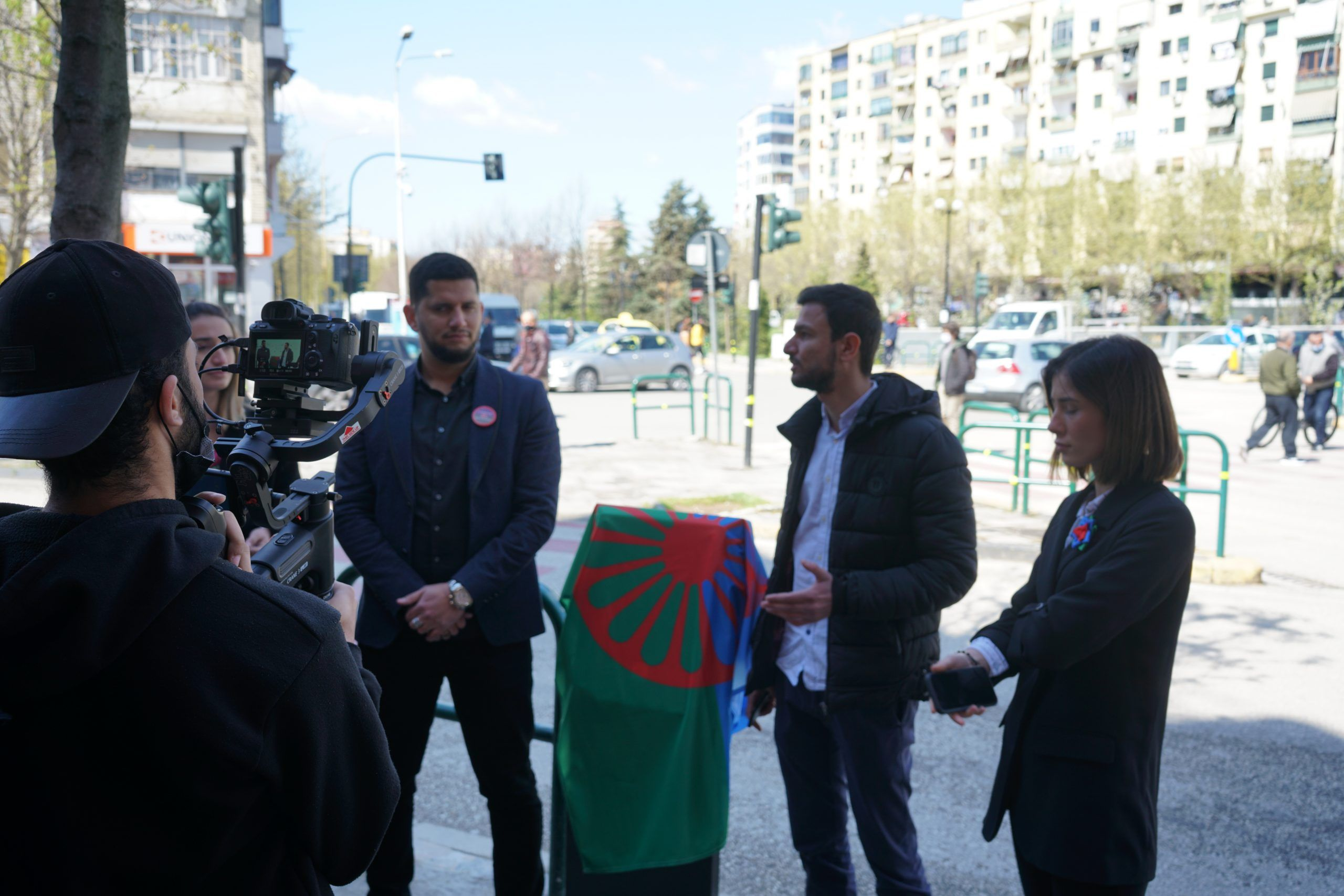 Speakers beside a plaque draped with the Romani flag at a public commemoration in Tirana
