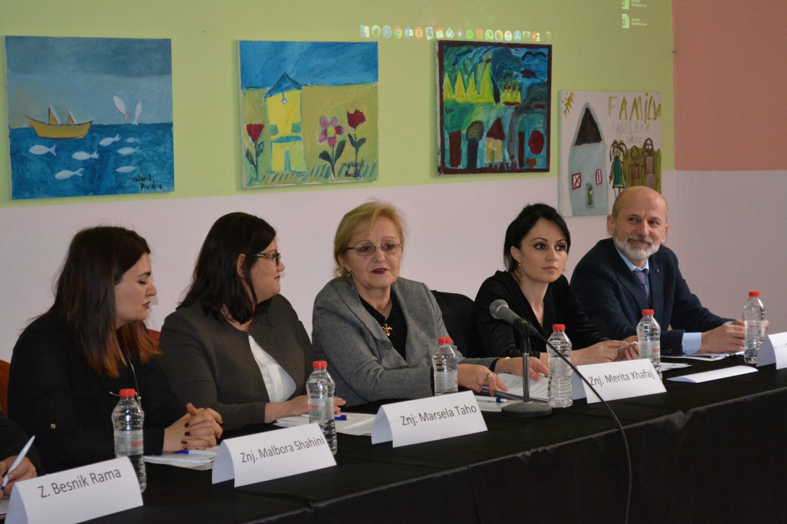 A panel of five speakers with name plates at a public roundtable held in a school